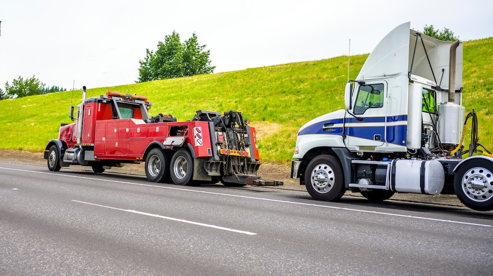 Powerful heavy-duty big rig mobile tow semi truck with emergency lights and towing equipment prepare to tow broken white big rig semi-truck standing out of service on the road side.